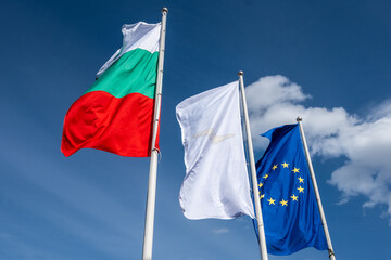 The Bulgarian flag alongside a white banner and the European Union flag prominently flutter on poles against a brilliant blue, cloudy expanse at the train station in Bansko, Bulgaria..