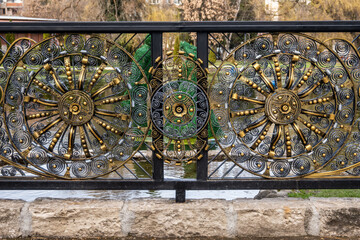 Intricate golden and dark metallic wrought iron fence with decorative wheel-like motifs and scrollwork, over water, showcasing detailed craftsmanship in Razlog, Bulgaria.