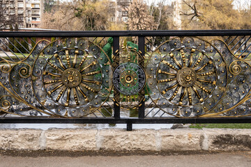 Wide shot of an elaborate golden and black wrought iron fence with intricate circular patterns and scrolls, situated in an urban park setting by water in Razlog, Bulgaria.