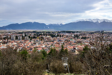Broad townscape of Razlog showcasing residential areas and modern blocks, framed by bare trees. Snow-covered Pirin Mountains loom under a dramatic cloudy atmosphere in early spring.