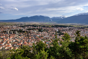 An expansive aerial panorama showcases Razlog town's densely packed red-roofed buildings, framed by vibrant green trees in the foreground, with majestic snowy mountains beyond.