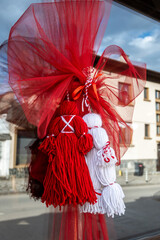 Traditional Bulgarian Martenitsa dolls, crafted from red and white yarn, hang inside a window for Baba Marta celebrations, reflecting an urban Razlog street scene.