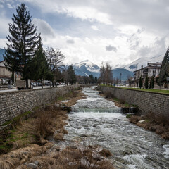 A river with small waterfalls flows through Bansko, Bulgaria, flanked by stone walls and leafless trees, leading towards snow-capped Pirin Mountains under a cloudy winter day.