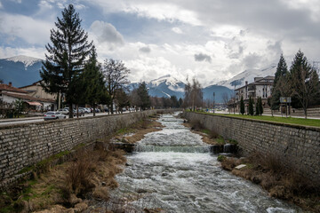 A picturesque river flows through Bansko, featuring stone embankments, leafless trees, and surrounding town buildings, set against the backdrop of snow-covered Pirin Mountains.