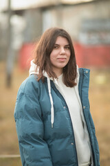Young woman with long dark hair wearing a blue puffer jacket and gray sweatpants is walking a dog along a rural path surrounded by tall grass and trees in the background
