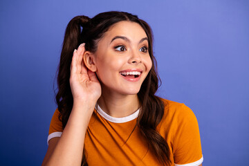 Young female teen with ponytails smiles excitedly while cupping her ear in an orange shirt against...