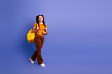 Young female student with a yellow backpack and book smiles as she walks against a purple violet backdrop