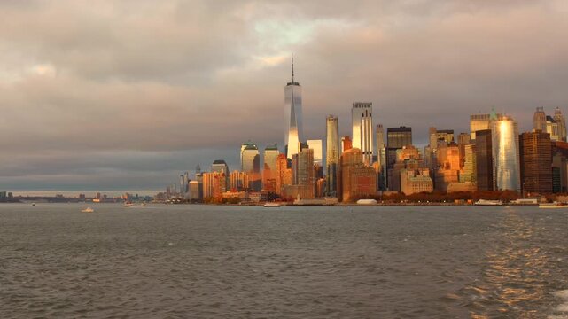 Wide shot of the Manhattan skyline during golden hour. The Freedom Tower and skyscrapers reflect warm sunlight across the water under a dramatic New York City sky.