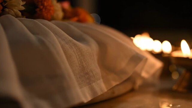 Close up Of White Fabric With Intricate Pattern Bordered By Dimly Lit Oil Lamps And Marigold Flowers In A Dark Ambience