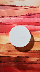 Blank white circular cosmetic product container rests atop vibrant swatches of shimmering bronze and deep red makeup textures for beauty flat lay.