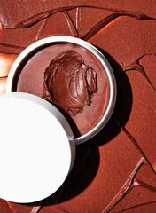 Rich dark brown creamy cosmetic product with metallic sheen presented in an open white jar against textured background