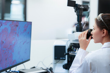 Side view of Asian female scientist looking through microscope near monitor. Researcher analyzing histology tissue sample in digital pathology lab for medical diagnosis.