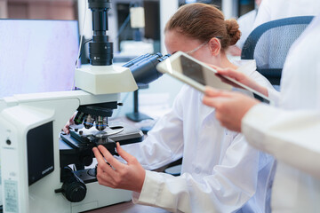 Team of female scientists collaborating in digital pathology lab. Young researcher adjusting microscope while colleague observes with tablet for medical diagnosis.