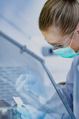 Close-up of focused female scientist wearing mask and safety glasses working in sterile fume hood. Researcher in blue gloves preparing tissue biopsy sample for histology analysis.