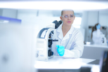 Caucasian female scientist using digital tablet to record data in modern laboratory. Researcher in white coat and blue gloves analyzing results near microscope.