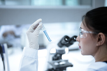 Female scientist holding test tube with clear liquid sample in modern lab. Researcher in white gloves inspecting chemical solution near microscope equipment.