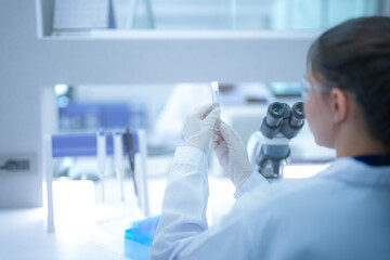 Focused female scientist holding a test tube to check the clear liquid sample in a modern sterile laboratory. Professional researcher analyzing chemical data for medical vaccine development.