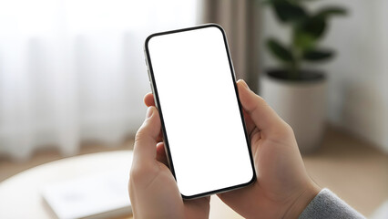 Hand holding mobile phone with blank white screen mockup in vertical portrait position in blurred home office interior for app design presentation digital marketing advertising media
