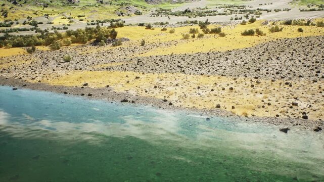A serene view of a riverbank featuring clear turquoise water, rocky shore, and patches of golden grass. Lush greenery rests in the background under a bright sky.