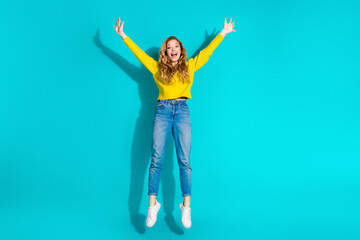 Young woman jumping with arms up wearing yellow sweater and jeans against bright blue background...