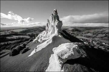 Rock structure rises sharply from grassy land in a countryside setting with distant hills and clouds