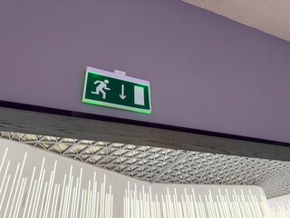 Sign on top of wall with navigational information showing direction to exit building in case of fire. Illuminated sign with running man symbol and arrow pointing toward door for emergency situation