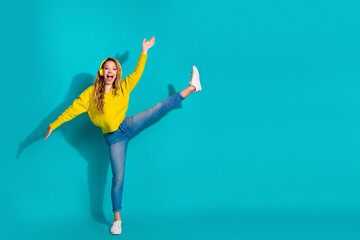 Young woman in bright yellow sweater jumps with one leg raised against a blue studio backdrop...