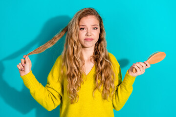 Young woman with long blonde hair in a bright yellow sweater holds a hairbrush against a blue...