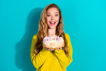 Young woman in a bright yellow sweater holds a colorful cake in front of a blue background smiling...