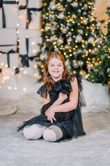 Young girl with long brown hair in a black dress, sitting on a soft carpet, holding a small black dog, surrounded by festive Christmas decorations and twinkling lights