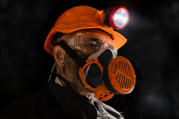 Coal miner with a dirty face in black dust, wearing a protective helmet with a flashlight, filtering respirator and work clothes for working in mine. Hard work underground in a coal mine, digging site