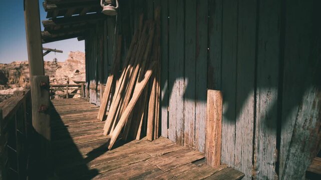 A rustic structure stands in the desert, its wooden porch bathed in sunlight. Stacked planks lean against the weathered wall, reflecting the tranquil yet rugged landscape.