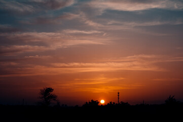 Obraz premium Bright setting sun amid layered orange-pink clouds, silhouetted rural horizon with tree and cell tower, serene evening landscape with warm atmospheric glow.