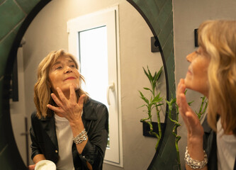 Senior woman doing her daily skincare routine applying cream to her face looking in mirror at home