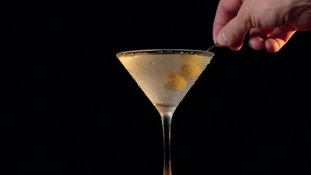 A person is dipping olives on a martini glass over a black background