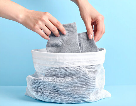 Hands placing grey delicate garments into white mesh laundry bag for washing on bright blue background.