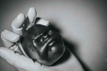 A black and white photograph of a shiny, sculptural voodoo doll head delicately held in the hand of a wooden mannequin. Soft shadows and eerie contrast enhance the surreal mood.