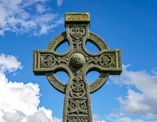 Celtic cross stands against blue sky with clouds in the background at historic graveyard during daytime