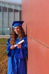 Happy young woman wearing graduation gown and cap holding her diploma on campus