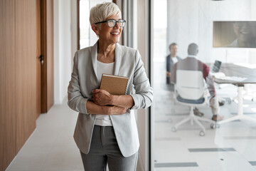 Professional Senior Woman In Office Hallway Smiling, Holding Notebook, Exuding Confidence And Leadership At Workplace