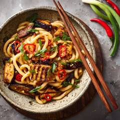 A bowl holds noodles topped with grilled vegetables, green onions, and chili. Chopsticks rest beside the bowl on a wooden surface. Spices are arranged around the dish.