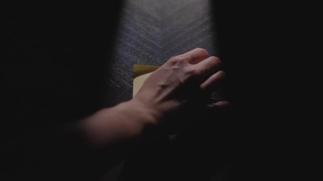 Slow motion close up footage of a hand writing out "Hola" on a paper note and placing it on a table. Shot in dramatic lighting for an impressive title or transition.