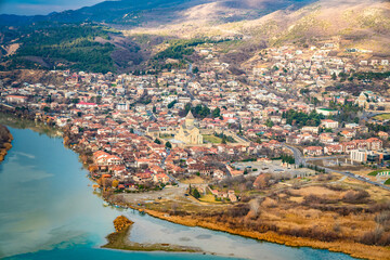From Jvari Monastery above Mtskheta, an elevated view looks over the old town clustered around Svetitskhoveli Cathedral beside the Mtkvari River, backed by rolling forested hills. © Auggie