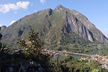 Panoramic autumn mountain landscape featuring a high peak view over a misty forest valley with green trees and a lake under a cloudy sky in Laos