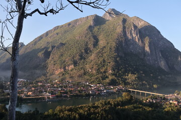 Panoramic autumn mountain landscape featuring a high peak view over a misty forest valley with green trees and a lake under a cloudy sky in Laos
