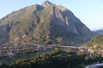Panoramic autumn mountain landscape featuring a high peak view over a misty forest valley with green trees and a lake under a cloudy sky in Laos