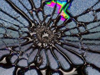Macro abstract of a cracked, reflective surface radiating from a center, featuring a bright dot pattern, dark fluid lines, and a patch of iridescent rainbow color.