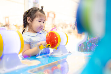 A cute little Asian girl is playing a coin-operated game machine in a shopping mall,Cute little girl adjusting knob and pressing red button while looking at screen of game machine at leisure center