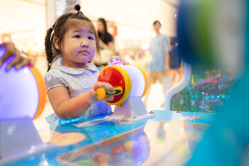 A cute little Asian girl is playing a coin-operated game machine in a shopping mall,Cute little girl adjusting knob and pressing red button while looking at screen of game machine at leisure center