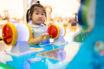 A cute little Asian girl is playing a coin-operated game machine in a shopping mall,Cute little girl adjusting knob and pressing red button while looking at screen of game machine at leisure center